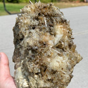 May include: A large, rough-hewn quartz crystal cluster with a mix of clear, tan, and brown hues. The crystal formation has many small, pointed crystal structures. The natural stone is held in a hand.