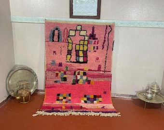 May include: A vibrant pink and red rug with a geometric design. The rug features a central pattern of colorful squares and rectangles, with a fringe along the bottom edge. The rug is displayed against a light-colored wall.