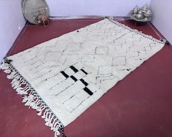 May include: Rectangular, off-white rug with a geometric pattern in black. The rug features a diamond and zig-zag design, with a section of solid black rectangles. Tassels line the edges. The rug is approximately 60 inches long.