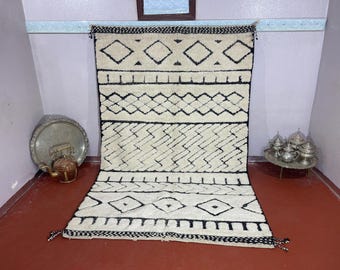 May include: Cream-colored, rectangular rug with a geometric black pattern. The design includes diamond, zigzag, and horizontal line motifs. The rug has a fringe on the ends and is displayed against a neutral wall.