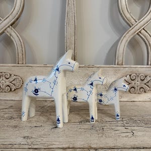May include: Three white wooden Dala horses of different sizes, decorated with blue floral patterns. They stand on a distressed wooden surface, with an ornate backdrop. These horses are a traditional Swedish folk art item.
