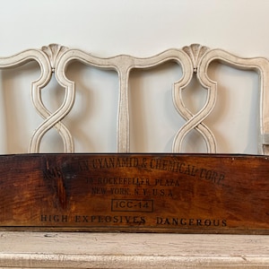 May include: A vintage wooden box with the text "AMERICAN CYANAMID & CHEMICAL CORP" and "HIGH EXPLOSIVES DANGEROUS" printed on it. The box is brown and sits on a white distressed chair.