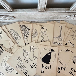May include: A collection of vintage flashcards with black line drawings on cream-colored paper. Illustrations include a crying face, a Native American headdress, a bunny, a scooter, a smiling sun, a bird, a ball, an apple, and a car.