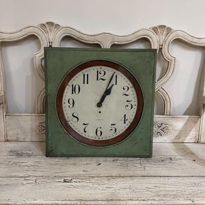 May include: Square, vintage-style clock with a green frame and a circular face. The clock face is cream-colored with black numerals and hands. The clock is set against a distressed white background, suggesting an antique aesthetic.