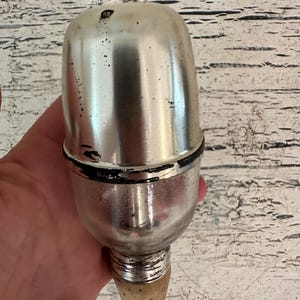 May include: A silver and black metal bottle stopper with a cork tip. The stopper has a round, bulbous top and a long, thin neck.