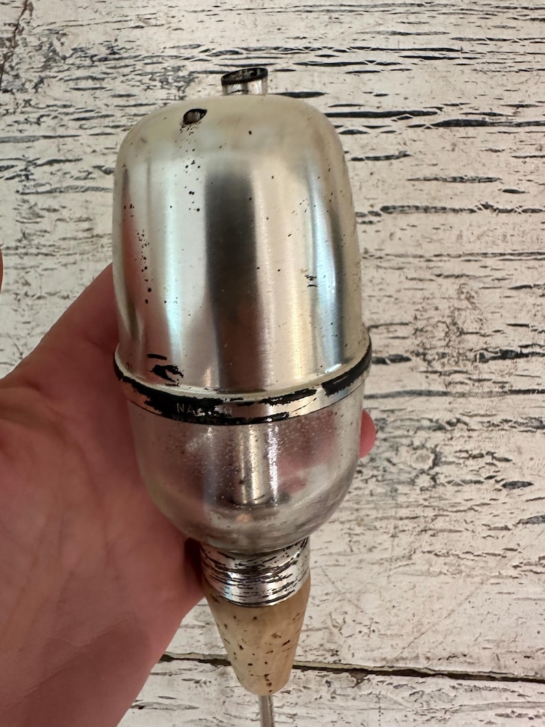 May include: A silver and black metal bottle stopper with a cork tip. The stopper has a round, bulbous top and a long, thin neck.