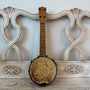May include: An antique banjo with a wooden neck and a round, aged drum. The drum has a light tan surface with dark brown metal trim. The banjo is set against a white, ornate background.