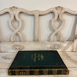 May include: A large, rectangular, dark green hardcover book titled Black's General Atlas of the World with gold lettering and a central emblem. The book features a dark leather-like spine with gold detailing and is resting on a white wooden bench.