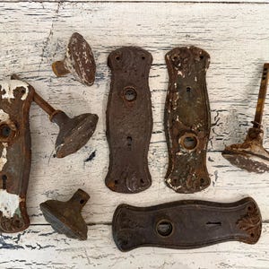 May include: A collection of antique metal door hardware including four rectangular backplates with keyholes and central openings, three round doorknobs, and one doorknob attached to a backplate. The metal surfaces show heavy rust and chipped white paint.