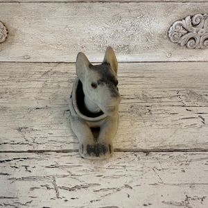 May include: A grey ceramic dog figurine with black markings. The figurine is sitting on a white wooden surface.