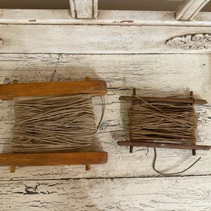 May include: Two rectangular wooden spools wrapped with tan-colored cord. The larger spool on the left features a thicker, lighter tan cord, while the smaller spool on the right is wound with a thinner, darker brown cord. Both spools have wooden crossbars.