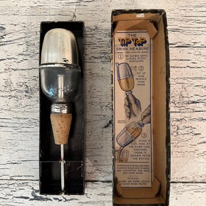 May include: Vintage Tip Top Drink Measure in its original box. The device is made of glass and metal and has a cork stopper. The box is brown with blue and white text and illustrations showing how to use the device.