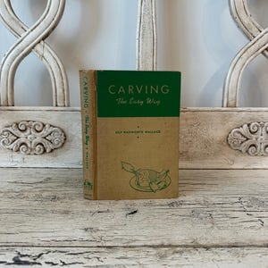 May include: A vintage book titled "CARVING The Easy Way" by Lily Haxworth Wallace. The book has a green and tan cover with illustrations of carved food. The book is displayed on a distressed white wooden surface.