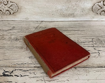 May include: A vintage book with a red cover and tan spine. The book's title is partially visible. The book is resting on a distressed, white-painted wooden surface, suggesting an antique or rustic aesthetic.