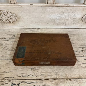 May include: A rectangular brown wooden cigar box with a hinged lid. The lid features the brand name Tilford in script, the text Worthy of the Name, and A Product of Park & Tilford. A green tax stamp is on the left side and a metal latch is centered.