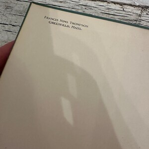 May include: A book page with the text "FRANCIS NIMS THOMPSON GREENFIELD, MASS." printed in black. The page is off-white with a green border. The background is a weathered, white-painted wooden surface.