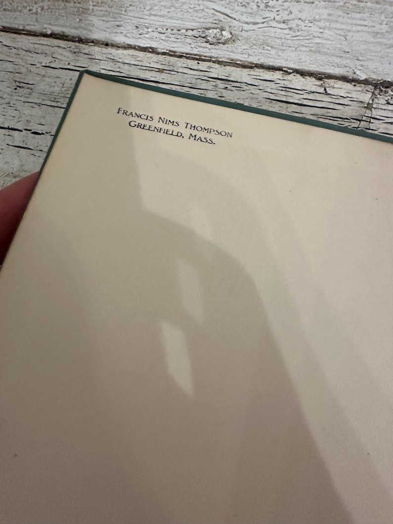 May include: A book page with the text "FRANCIS NIMS THOMPSON GREENFIELD, MASS." printed in black. The page is off-white with a green border. The background is a weathered, white-painted wooden surface.