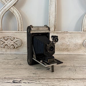 May include: A vintage black folding camera with a dark accordion-style bellows, a metal lens housing, and a small viewfinder. The front plate features the text No. 1 Autographic Kodak Jr. and a small metal stand extending from the base.