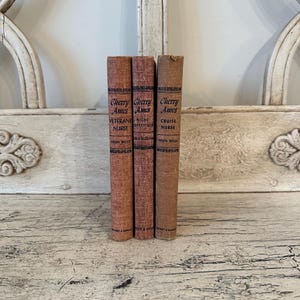May include: Three vintage books with worn, textured covers. The spines display the title "Cherry Ames" in a decorative font, along with the titles "Veterans Nurse", "Night Supervisor", and "Cruise Nurse". The books are in shades of brown and red.