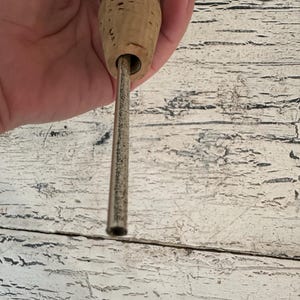 May include: A close-up of a cork stopper with a metal tube inserted through the center. The cork is brown and the tube is silver.