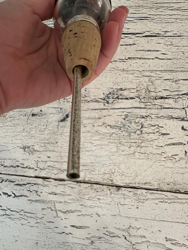 May include: A close-up of a cork stopper with a metal tube inserted through the center. The cork is brown and the tube is silver.