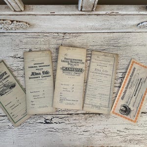 May include: A collection of vintage insurance documents, including policies and certificates, are arranged on a distressed white wooden surface. The documents feature text and graphics, with the "Etna Life Insurance Company" logo visible on several items.