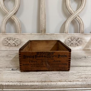May include: A vintage wooden box with the text "HIGGINS Drawing Board & Library Paste" printed on the side. The box is rectangular and appears to be made of aged wood. The box is sitting on a distressed white surface.