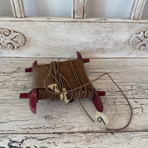May include: A vintage wooden spool with brown thread and red ends. The spool has small paper tags with numbers. The background is a distressed white wooden surface.
