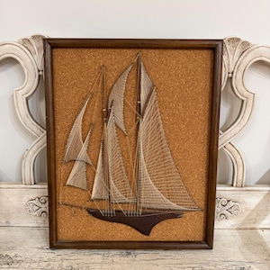 May include: A framed string art piece featuring a sailboat design. The artwork uses thin beige thread stretched across a textured cork background to form the sails and rigging, mounted on a dark brown wooden hull, all enclosed in a dark wood frame.