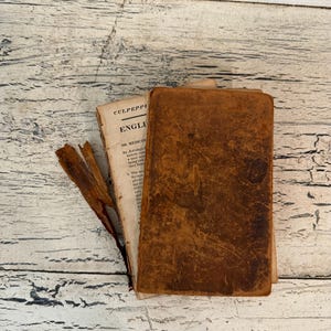 May include: An antique book with a worn, dark brown leather cover resting on top of an open page from Culpeper's English Physician. The exposed page features black printed text and a small, dried, brown botanical specimen placed to the left.