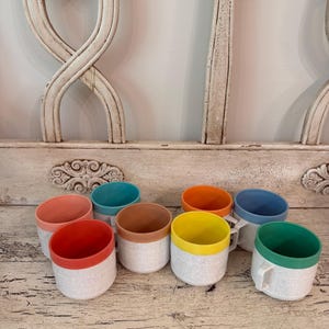 May include: A collection of eight ceramic mugs with speckled white bodies and solid colored rims in shades of coral, teal, terracotta, orange, yellow, blue, and green. Each mug features a simple cylindrical shape and a small side handle.