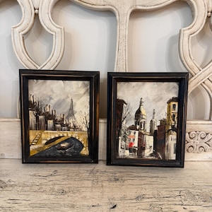 May include: Two framed oil paintings depicting Parisian cityscapes. The left painting shows the Eiffel Tower and a river with a boat. The right painting features a street scene with a domed building. Both artworks are in dark, distressed frames.
