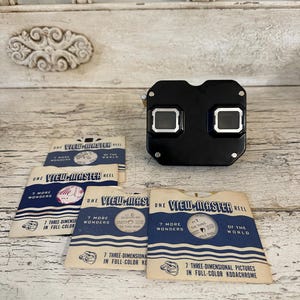 May include: A black View-Master viewer with silver accents and several View-Master reel packets. The packets are blue and white, with text that reads "7 More Wonders" and "7 Three-Dimensional Pictures in Full-Color Kodachrome."
