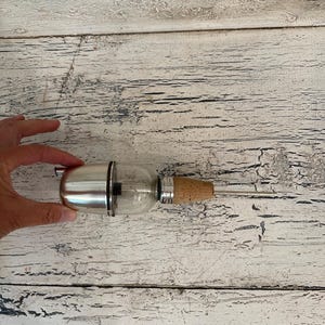 May include: A silver and glass bottle stopper with a cork top. The bottle stopper is on a white wooden surface.