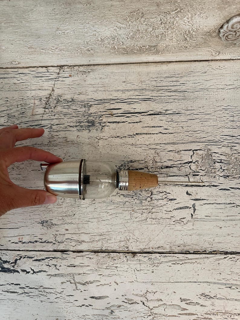 May include: A silver and glass bottle stopper with a cork top. The bottle stopper is on a white wooden surface.