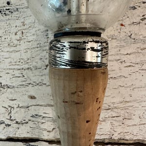 May include: A glass bottle stopper with a silver metal top and a cork base. The cork is brown and has a textured surface.