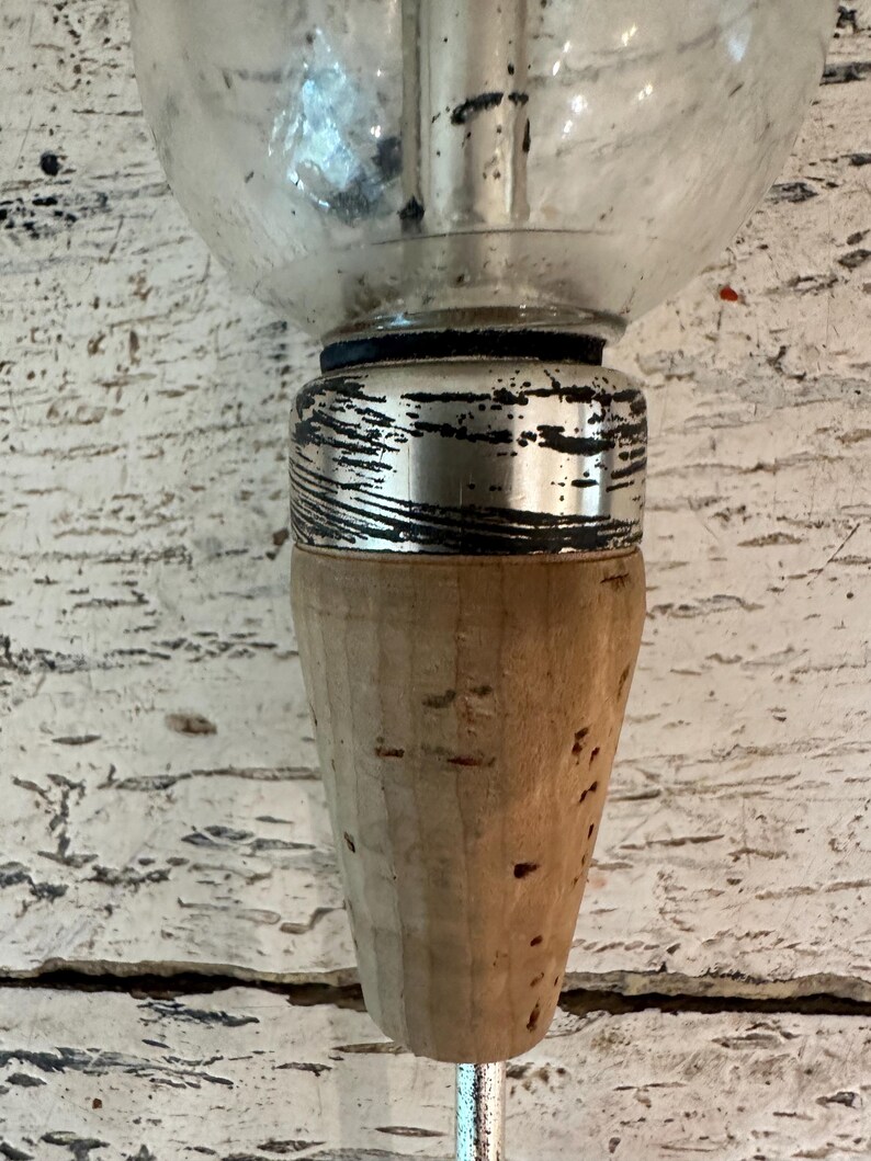 May include: A glass bottle stopper with a silver metal top and a cork base. The cork is brown and has a textured surface.