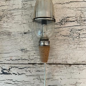May include: A silver and clear glass bottle stopper with a cork and a long metal needle. The stopper is designed to be inserted into a bottle of wine or other beverage to keep it fresh.