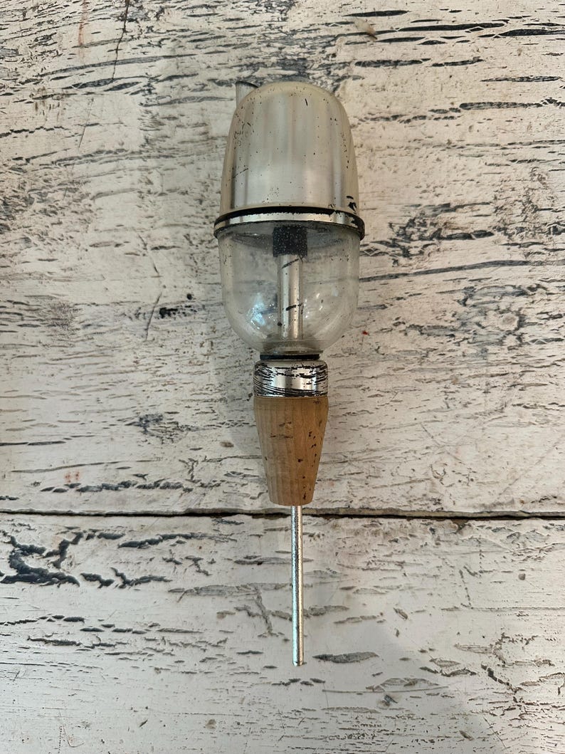 May include: A silver and clear glass bottle stopper with a cork and a long metal needle. The stopper is designed to be inserted into a bottle of wine or other beverage to keep it fresh.
