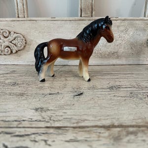 May include: A ceramic horse figurine with a glossy finish. The horse has a brown body, black mane and tail, and white legs. A small label on the side reads "Toby -116". The figurine is posed on a wooden surface.