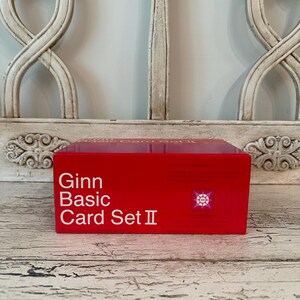 May include: A red box of Ginn Basic Card Set II. The text is in white, with a small graphic on the right side. The box is sitting on a distressed, white-painted wooden surface.