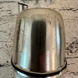 May include: A silver metal bottle stopper with a spout. The stopper has a black rubber seal around the top.