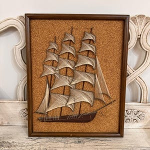 May include: A framed string art piece featuring a sailing ship with multiple masts and white thread sails. The ship is set against a textured brown cork background and enclosed in a dark wooden frame, creating a detailed nautical wall hanging.