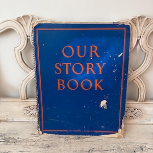 May include: A rectangular spiral-bound book with a dark blue cover featuring the orange text "OUR STORY BOOK" and "EASY GROWTH IN READING" at the bottom. The cover shows signs of wear, including scuffs, scratches, and frayed edges.