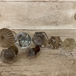 May include: A collection of eight vintage glass door knobs in various shapes and sizes. The knobs are clear glass and have a slightly frosted appearance. They are arranged on a light wood surface.