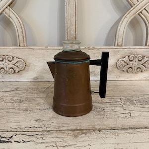 May include: A small, copper-colored coffee pot with a black handle and a glass top. The pot has a spout and a lid. The pot is sitting on a distressed, white-painted wooden surface.