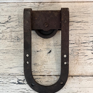 May include: A rustic, U-shaped metal barn door roller with a wheel. The roller is dark brown with visible signs of wear and tear, suggesting age and use. The design includes a top plate and multiple mounting holes.