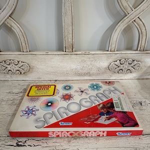 May include: A vintage Spirograph drawing toy set. The box is white with red accents and features the word "Spirograph" in large, bold letters. The box also includes colourful geometric designs and the Kenner logo. The box is on a white, distressed wooden surface.