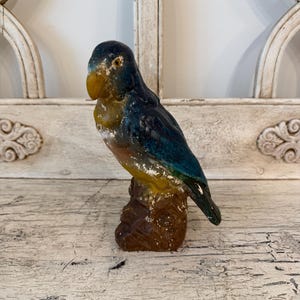 May include: A vintage-style parrot figurine in shades of blue, yellow, and brown. The bird is perched on a brown base, with a detailed, aged appearance. The figurine is likely made of ceramic or a similar material.
