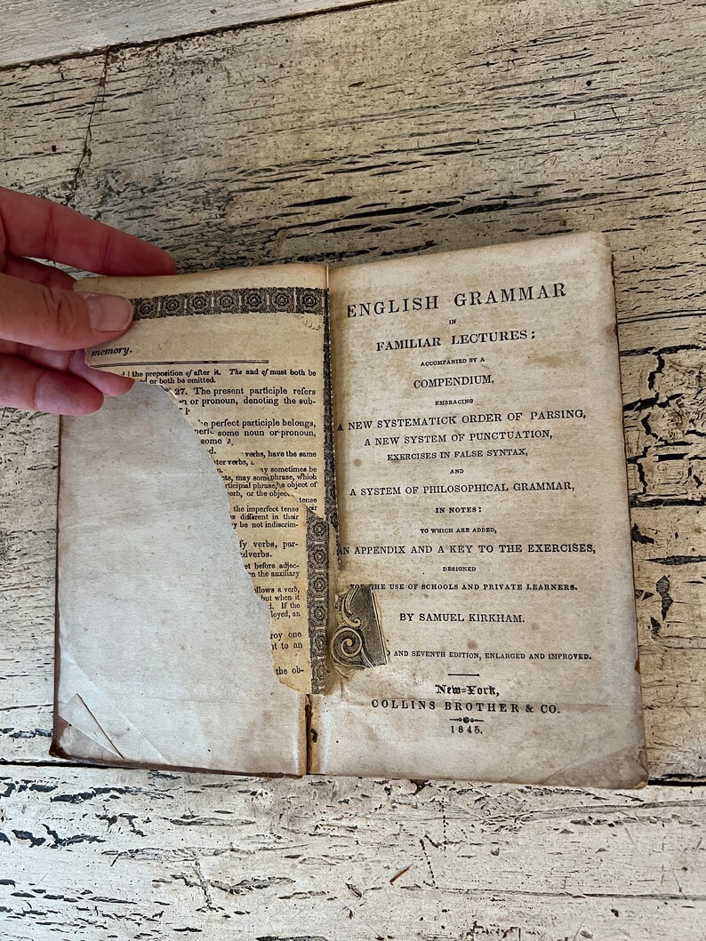 May include: An antique book titled "English Grammar in Familiar Lectures: Accompanied by a Compendium" by Samuel Kirkham. The book is open to the title page and the copyright page, which states that it is the seventh edition, enlarged and improved, published in New York by Collins Brother & Co. in 1845.